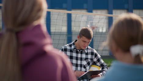 coach with volleyball under arm instructing new student in outdoor volleyball court while holding clipboard, preparing for practice, background features net and buildings