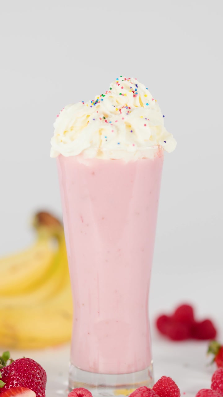 Granella colorata cade su un frullato di fragole e banane con panna montata, circondato da frutta fresca, con luce intensa.