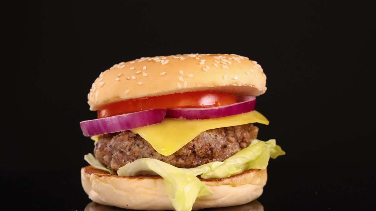 A juicy beef burger with cheese, lettuce, tomato, and onion is gradually zoomed out against a clean black backdrop with studio lighting