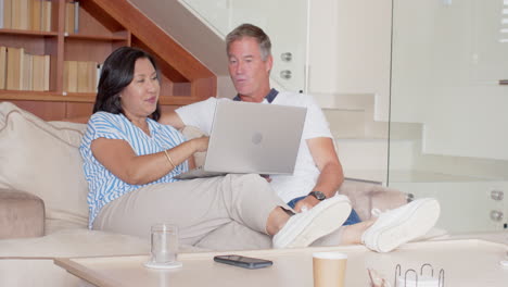 senior diverse couple relaxing on sofa, using laptop and enjoying time together at home