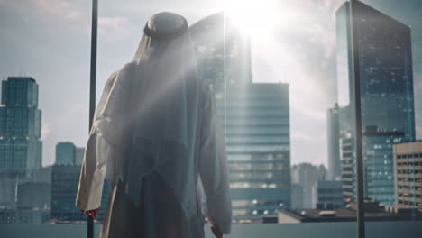 successful muslim businessman in traditional white kandura walking in his modern office looking out of the window on big city with skyscrapers. successful saudi, emirati, arab businessman concept.