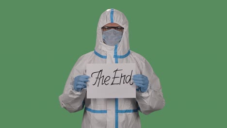 portrait of a man in a protective suit, goggles, gloves and a respirator looking at the camera holding a the end sign. green screen, chroma key. coronavirus death concept. slow motion. close up