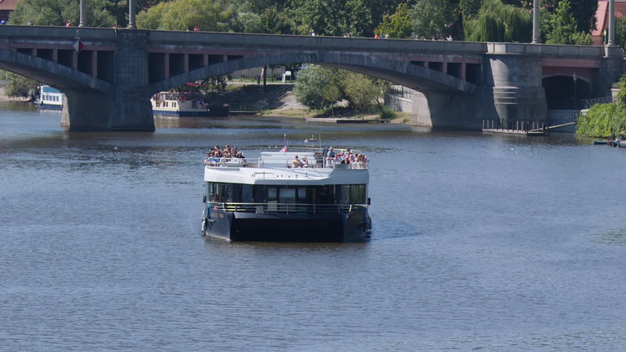 Passenger boat moves toward stone bridge on Vltava River, sunny summer day, steady camera