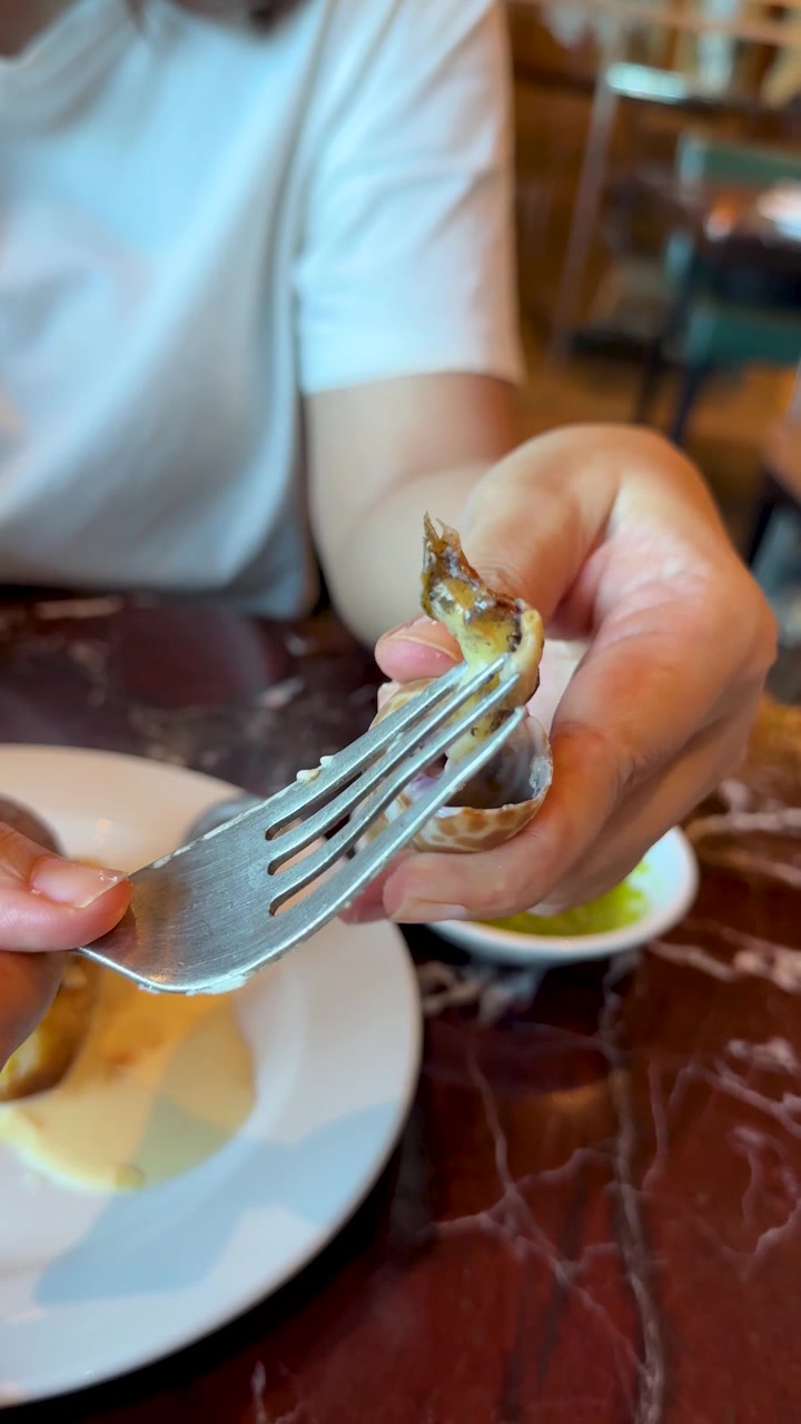 A person uses a fork to remove meat from a grilled spotted babylon snail shell