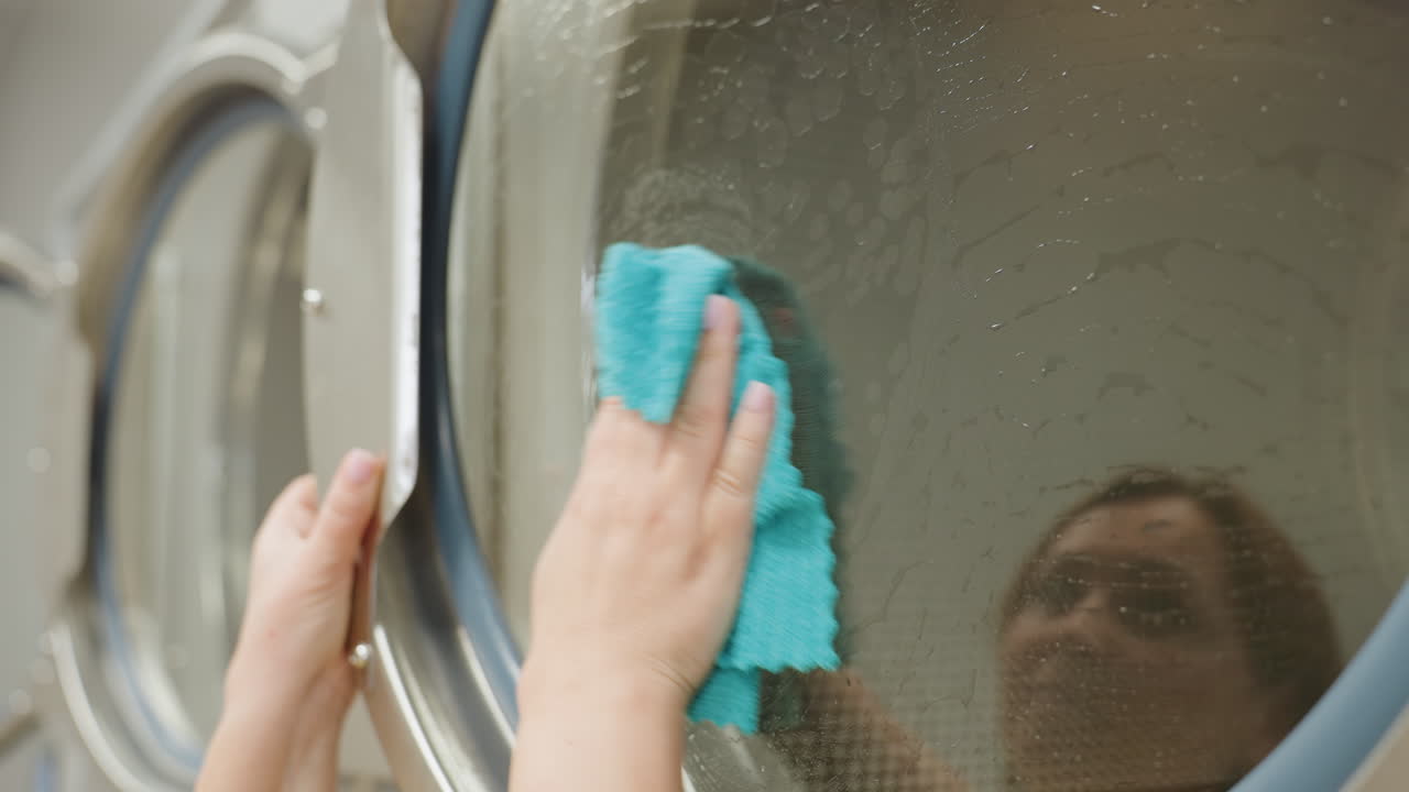 Close up business woman carefully dusting transparent door of washing machine with soapy towel, reflection visible on glass surface as she thoroughly wipes during cleaning process