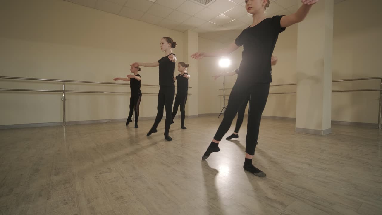 группа молодых студентов балета в черной танцевальной одежде практикует позиции в просторной балетной студии с деревянным полом и настенными решетками. сосредоточенные выражения и синхронизированные движения.