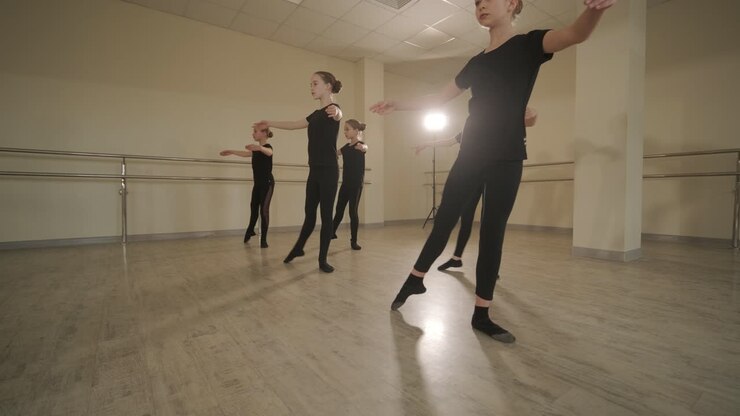 Un gruppo di giovani allievi di danza classica in abbigliamento da ballo nero che eseguono esercizi in un ampio studio di danza con pavimenti in legno e barre a muro. Espressioni concentrate e movimenti sincronizzati