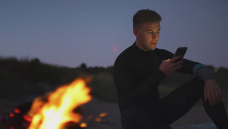 Mann-Sitzt-Auf-Einem-Surfbrett-Am-Lagerfeuer-Am-Strand-Und-Benutzt-Sein-Mobiltelefon,-Während-Die-Sonne-Hinter-Ihr-Untergeht