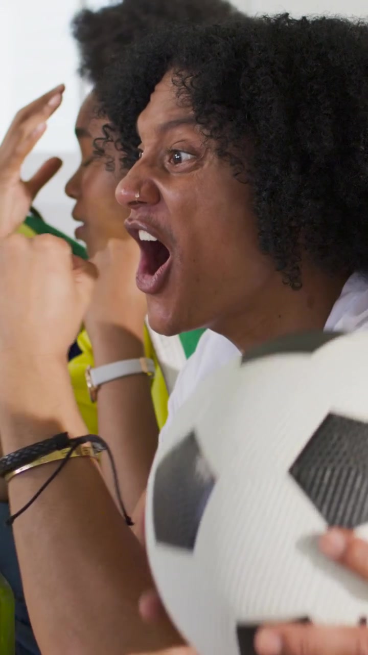 Video vertical: Aficionados gritando con camisetas, haciendo muecas en el sofá, reaccionando a un partido en la televisión con un balón de fútbol