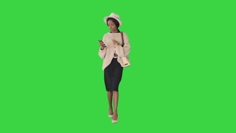 stylish african american woman in knitwear and hat using her phone while walking on a green screen, chroma key