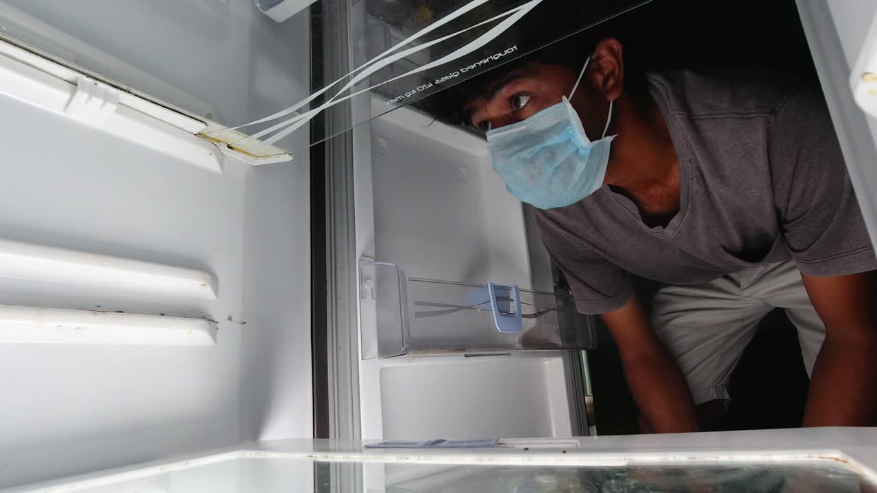 hombre con una máscara médica mira en el refrigerador o refrigerador vacío para comida - concepto de que no hay comida en la despensa disponible durante la cuarentena en casa en covid-19 o coronavirus