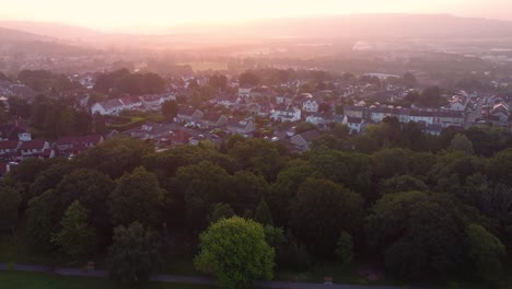 pushing aerial towards houses at dawn with hazy sunrise covering valley with nearby park with trees. drone view of densely populated estates and streets