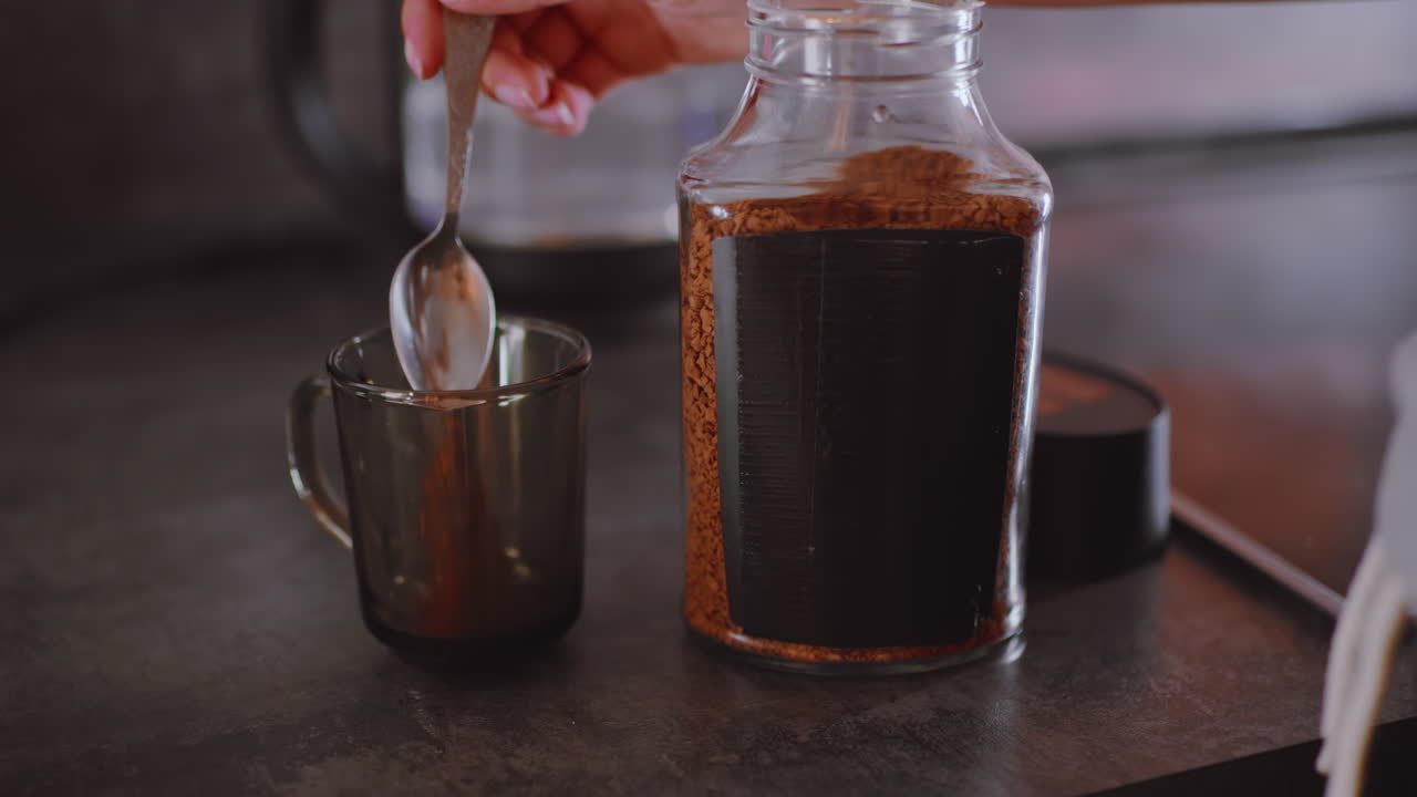 Nærbillede af elkedel med vand, der varmer, mens en hånd rækker ud efter låget, lige før personen hælder kaffe i koppen, og der dannes damp, mens forberedelsen af den varme drik begynder i køkkenet