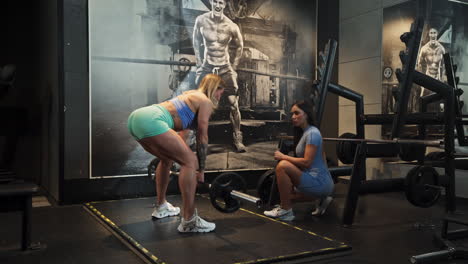 caucasian blond woman doing deadlifts at the gym while brown girl is watching, indoor, wide shot