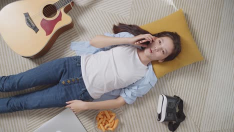 vista superior de una adolescente asiática hablando en su teléfono inteligente mientras yace en la alfombra en el suelo de su casa