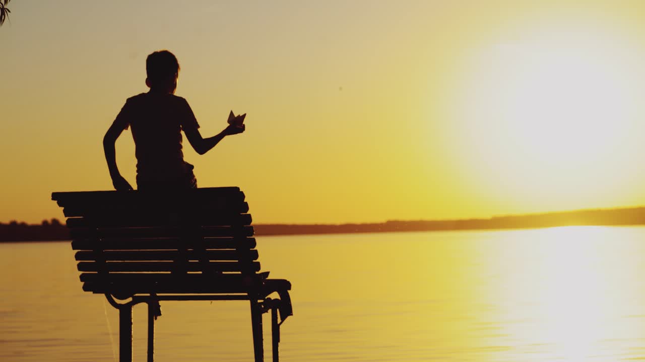 Little boy is playing with a paper boat on an old bridge by the river. Silhouette of little boy sitting alone at the river on sunset background