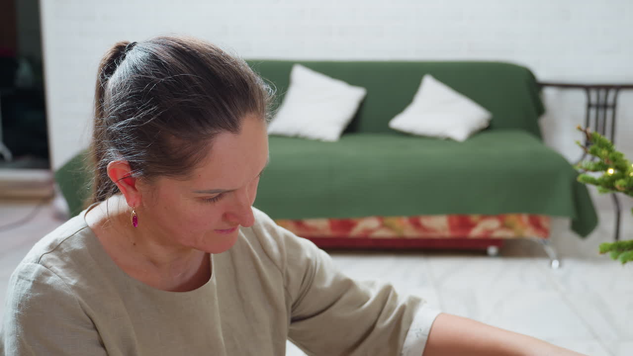 Woman in casual gown close-up view observing Christmas tree with soft background showing green couch and festive decorations, creating cozy peaceful indoor atmosphere during joyful holiday season