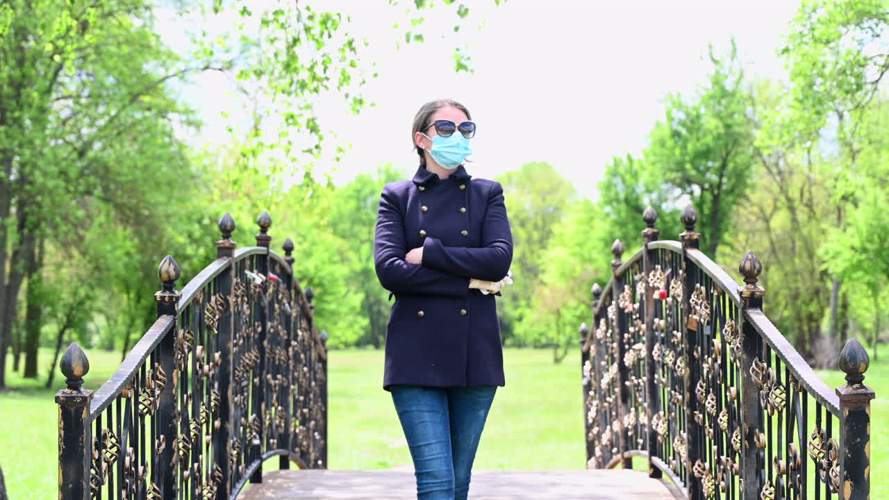 A woman wearing a mask walks across a decorative iron bridge surrounded by greenery on a bright spring day