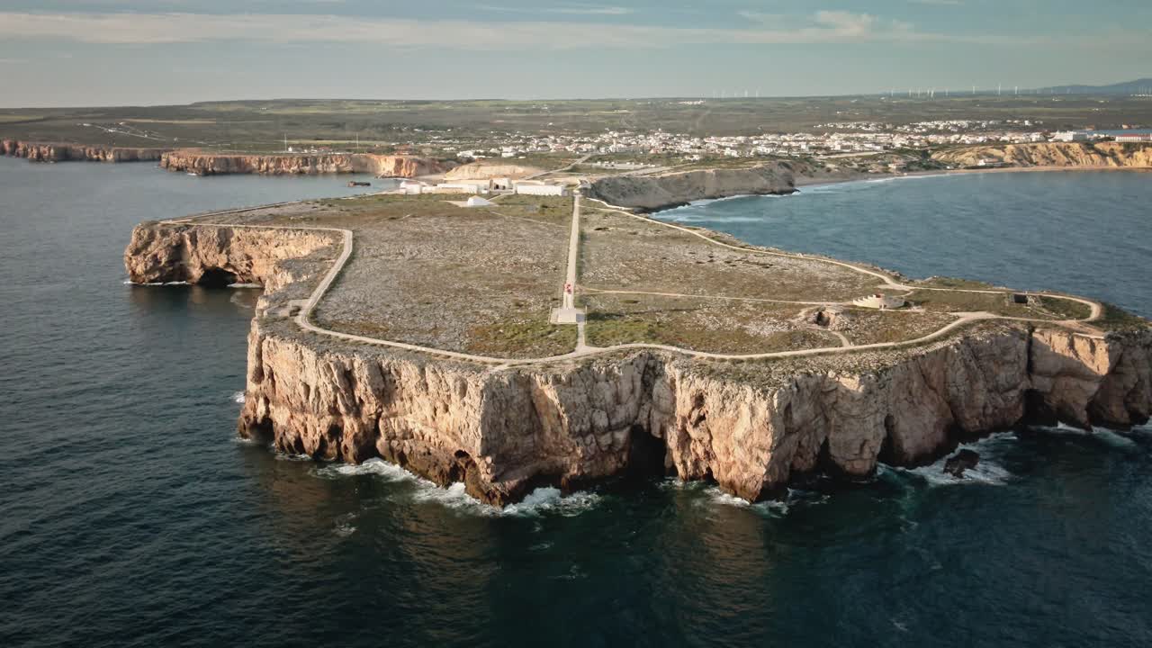 Cinematic aerial shot drifting backward to unveil the dramatic cliffs, fort, and lighthouse at Sagres—the southwestern tip of Portugal, surrounded by ocean