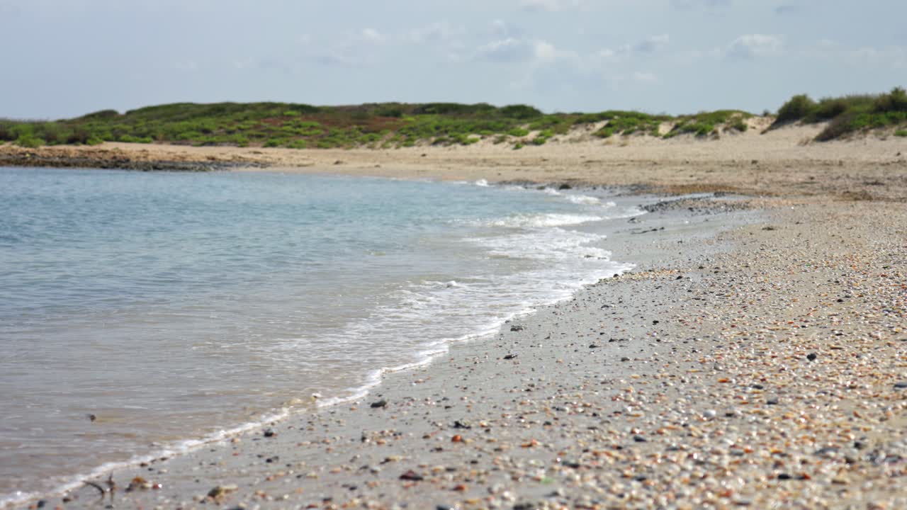 Gentle ocean waves wash over a pebble-covered beach with green coastal hills in the background under a sunny sky.