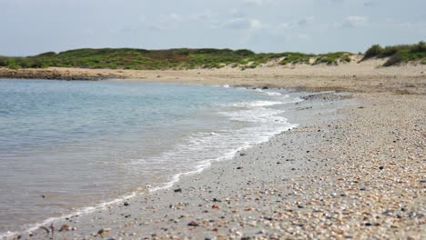 gentle ocean waves wash over a pebble-covered beach with green coastal hills in the background under a sunny sky.