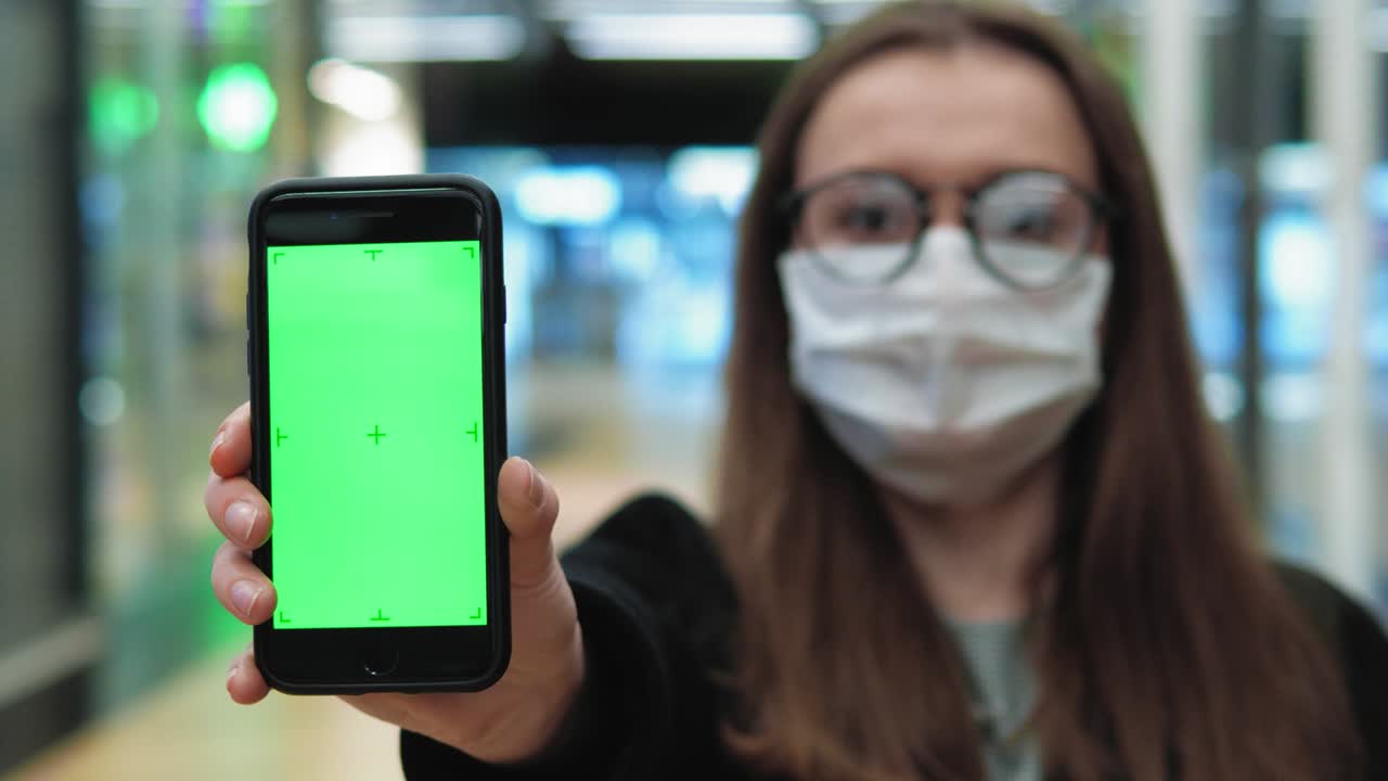 mujer joven de pie con una máscara médica y gafas. sostener el teléfono con pantalla verde. mujer mirando a la cámara. cuidado de la salud y concepto médico de coronavirus. retrato en primer plano en 4k uhd 2160p
