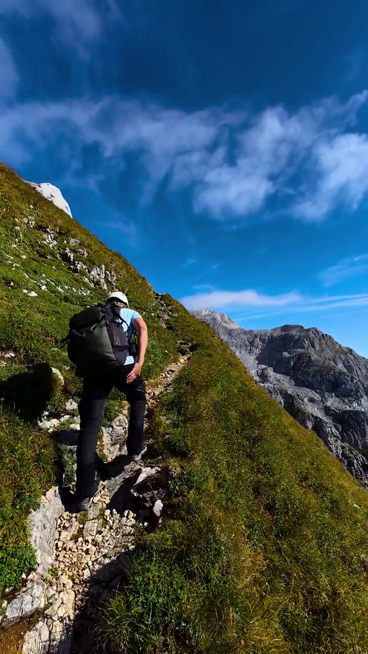 Vertical handheld view, male hiker ascending a steep mountain trail near Bovški Gamsovec in Slovenia, wearing a safety helmet and backpack in the dramatic Julian Alps