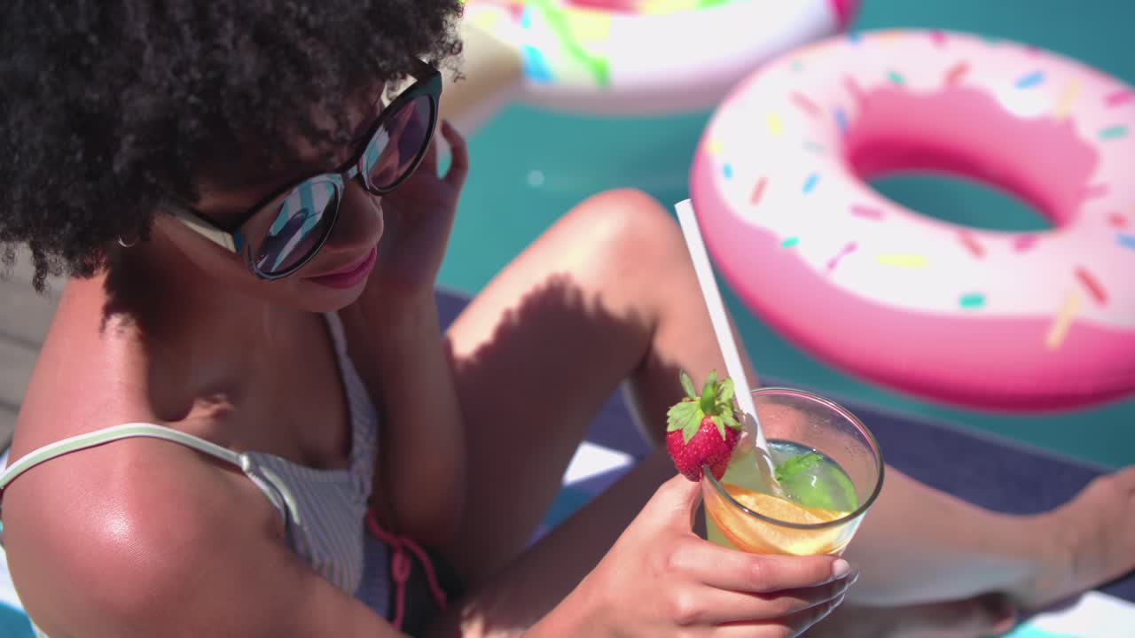 A câmera se move mostrando uma mulher mexendo um coquetel, bebendo com um canudo enquanto relaxa na piscina, perto de uma boia rosa.