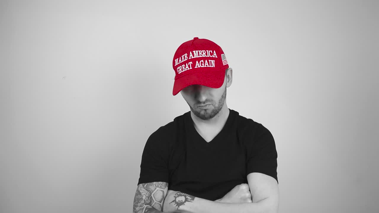Grayscale footage of a tattooed man with short hair, beard and a black shirt putting on a vibrant red MAGA or Make America Great Again Trump presidential merch hat, crossing his arms and nodding happy