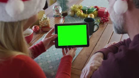 Pareja-Caucásica-Sonriente-Con-Sombreros-De-Papá-Noel-Haciendo-Videollamada-Navideña-Con-Tableta,-Con-Pantalla-Verde