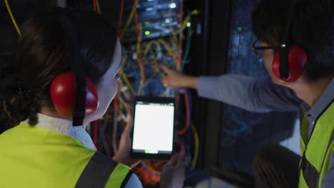 Diverse-female-and-male-it-technicians-wearing-headphones-using-tablet-checking-computer-server