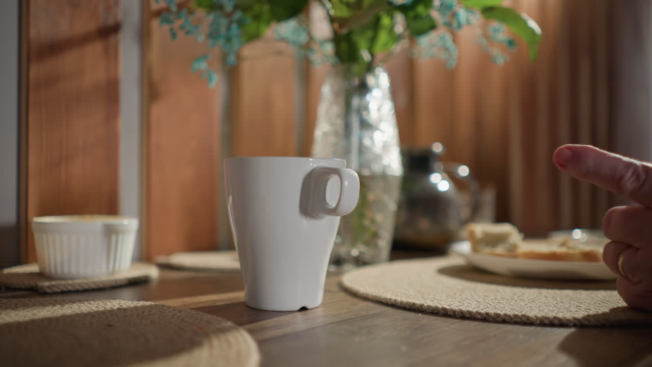 Close-up of white coffee cup on table with decorative flowers and food on a plate. Bright natural light, cozy indoor setting with soft textures, warm atmosphere, calm moment captured