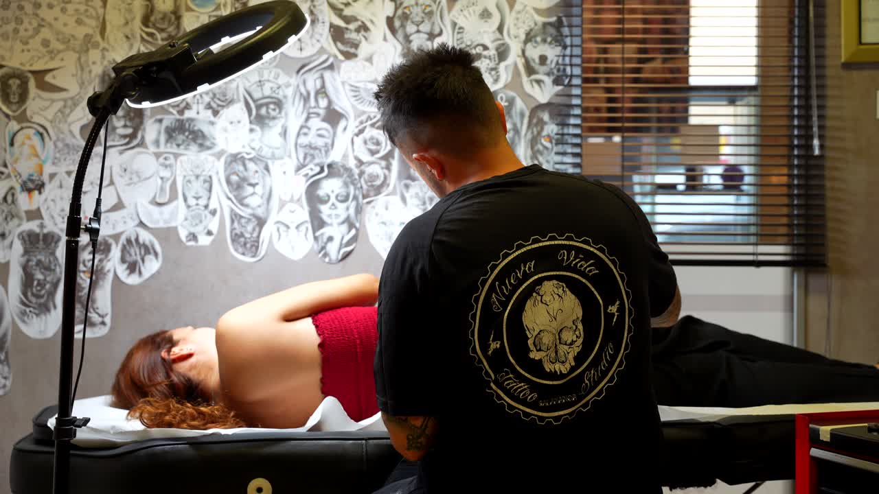 Rear shot of a tattoo artist taking some ink and tattooing a woman lying on a stretcher