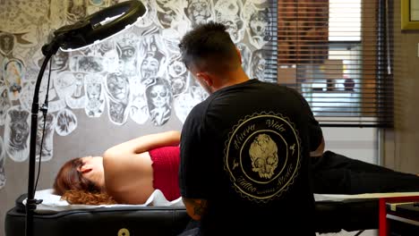 rear shot of a tattoo artist taking some ink and tattooing a woman lying on a stretcher