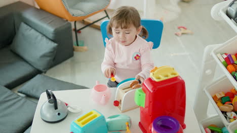 Niña-Finge-Comer-Con-Una-Cuchara-De-Juguete-De-Un-Recipiente-Vacío-De-Plástico-En-La-Cocina-Sentada-Junto-A-La-Mesa-En-La-Sala-De-Juegos-De-Casa