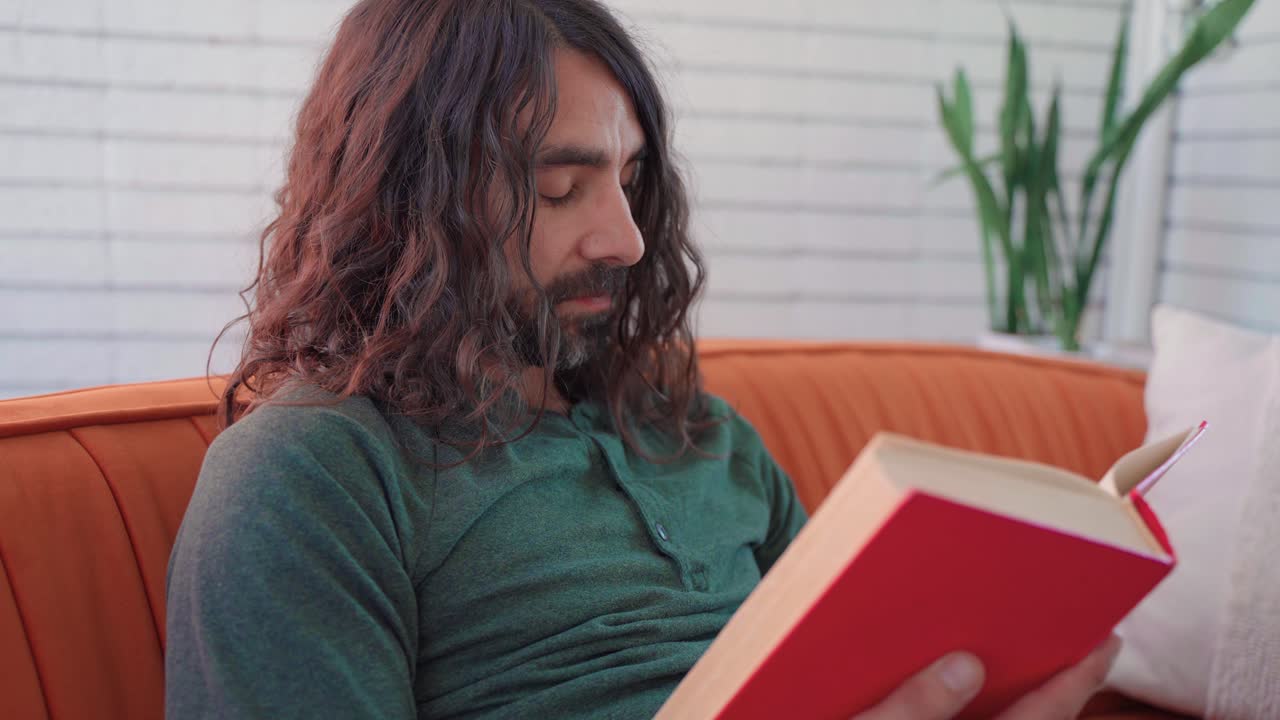 hombre hispano con cabello largo y castaño rizado y barba con camisa verde leyendo tranquilamente un libro rojo en un sofá naranja en una habitación blanca