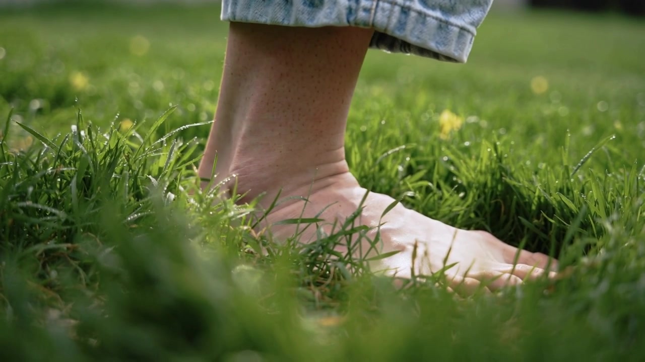 Contacting short grass, bare foot shifting weight on lawn for balance, rolled denim, yellow flowers