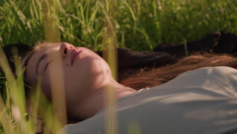 close-up of young woman lying in a grassy field, thoughtfully gazing up, immersed in nature, surrounded by tall, sunlit grass that softly frames her face