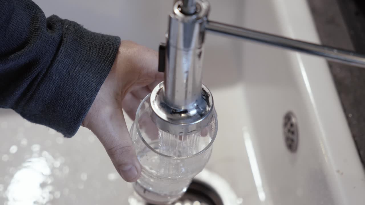 Overhead shot of a person filling a clear glass with tap water from a sink, symbolizing hydration and daily routine, dominant clear and metallic tones