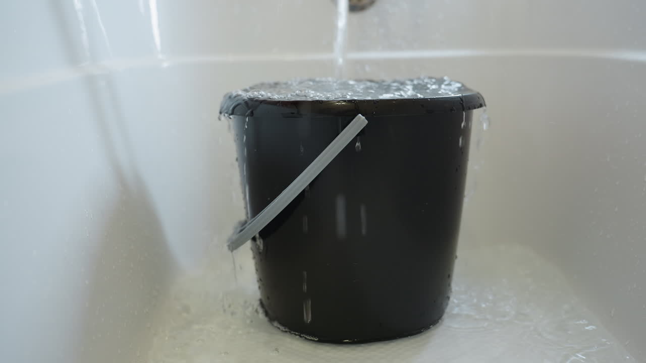 Water pouring into black bucket placed in bathroom sink, overflowing as liquid spills down sides, handle visible on container, concept of excess and cleaning preparation