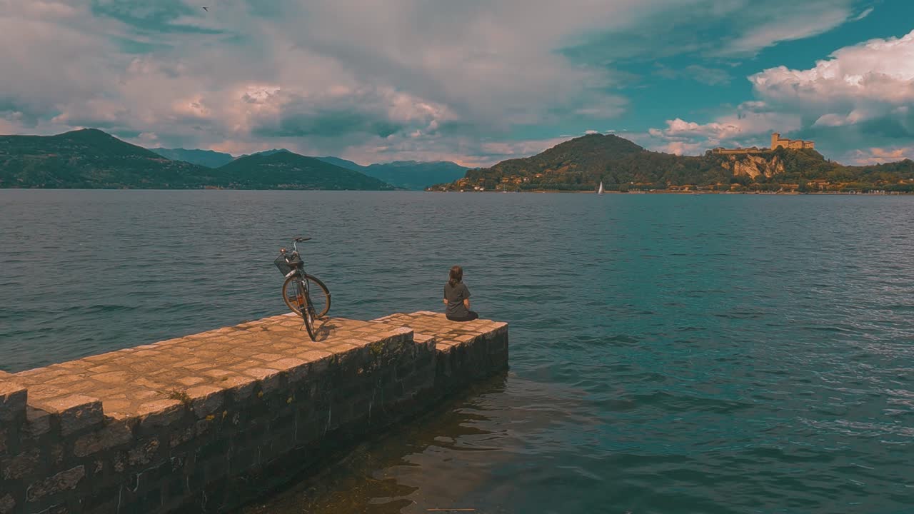 Veduta di schiena di donna solitaria con bicicletta dietro, seduta sul bordo di un molo sul Lago Maggiore, Italia