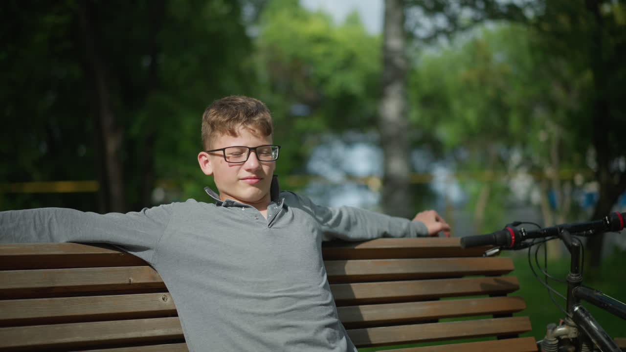 젊은이는 나무 벤치에 앉아서 팔을 넓게 펴고, 주차된 자전거의 손잡이가 에 보이고, 흐릿한 사람들이 나무와 초록색으로 배경에서 고 있습니다.