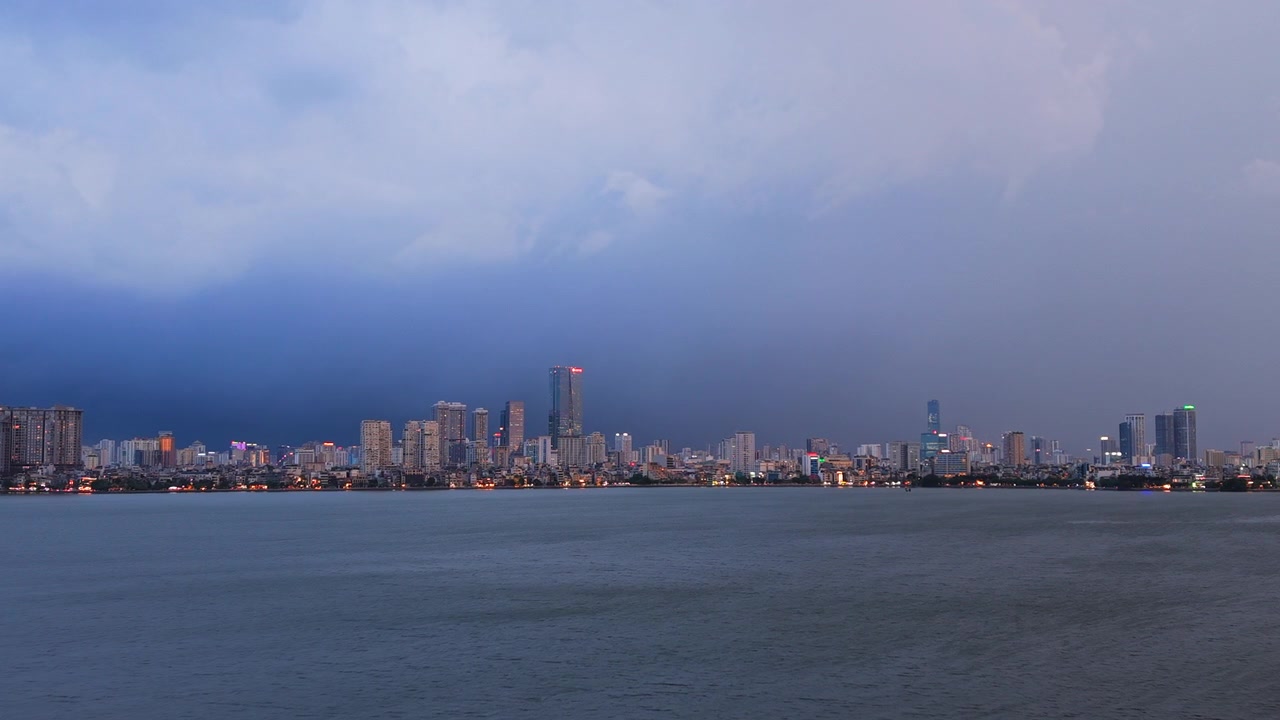 Ampia inquadratura cinematografica drammatica di un temporale con fulmini che si manifesta sullo skyline di Hanoi e sul Lago Tay Ho Ovest durante l'ora blu. Ottimo per notizie meteo e documentari di viaggio
