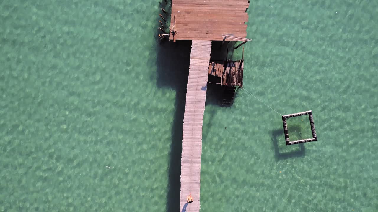 Aerial, birdseye, drone shot, over a asian woman walking on a wooden pier, surrounded by turquoise, shallow water, on a sunny day, at Koh Kood island, in Thailand