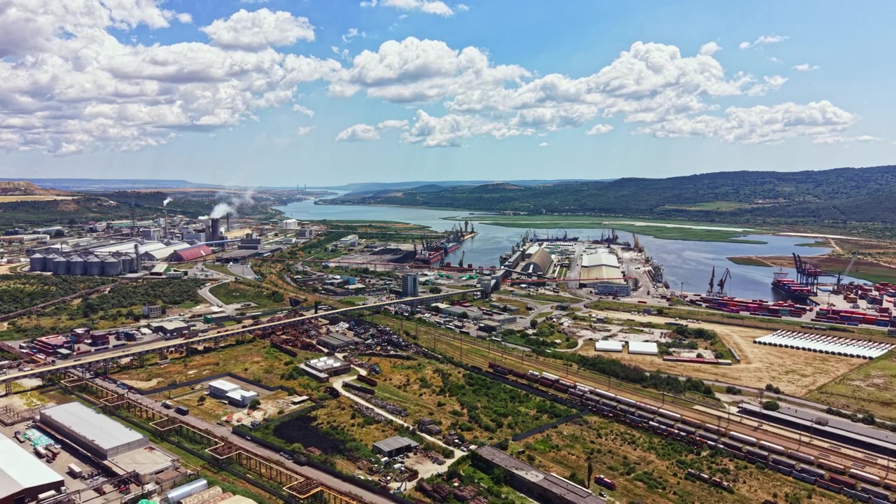 Area industriale vicino a un grande specchio d'acqua con porti, fabbriche e vegetazione, osservata di giorno sotto un cielo azzurro con nuvole.