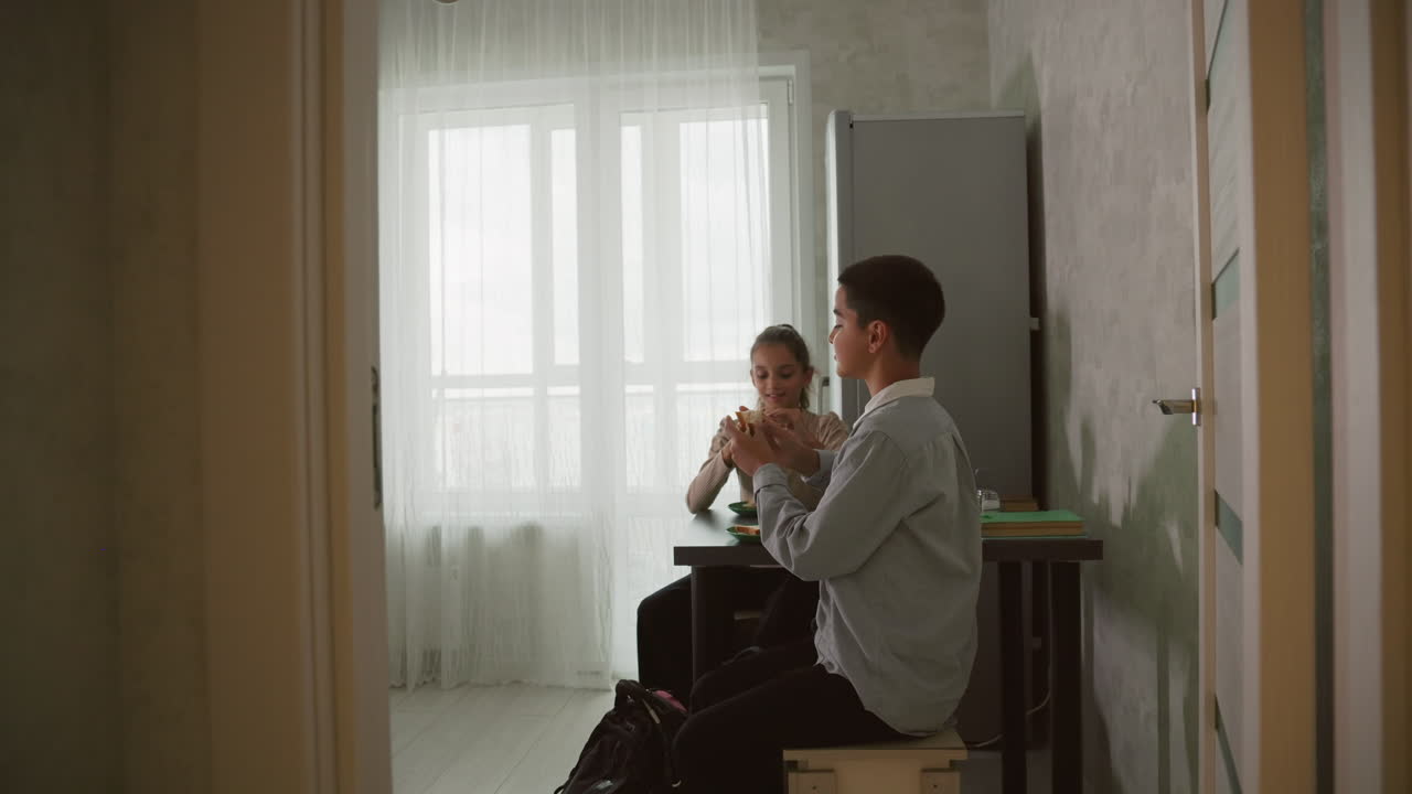 woman in pink robe serving sandwiches on plates to children at kitchen table, one child eagerly reaching for sandwich while other looks bored, bright morning light in modern home kitchen