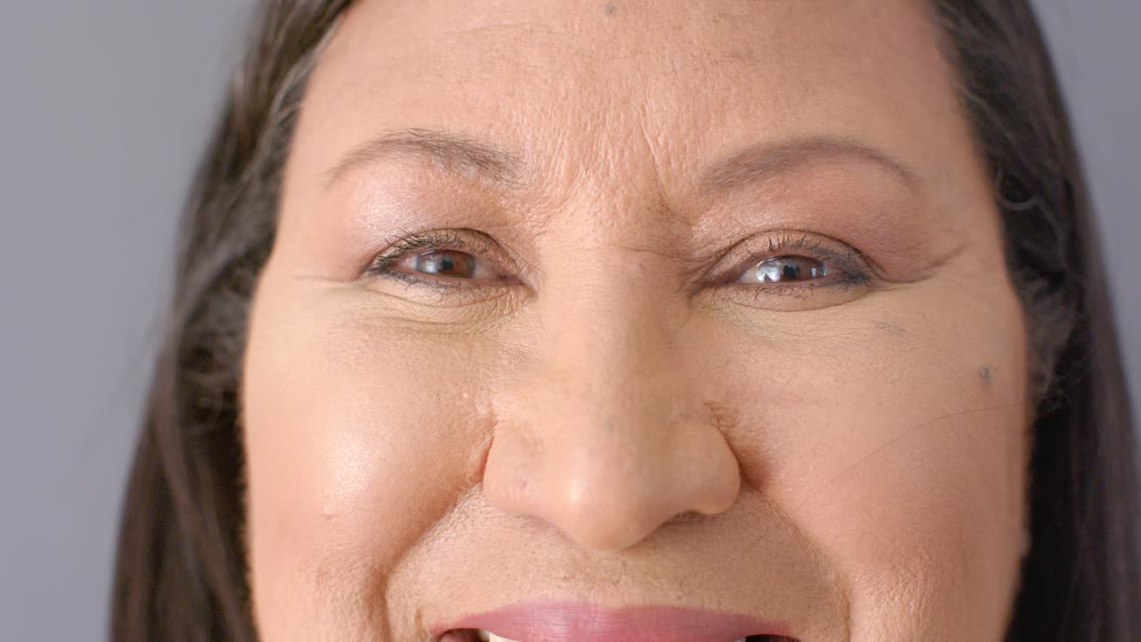 Oudere vrouw neemt een aanwijzing, lacht en toont haar tanden, met eyeliner en lippenstift op een grijze achtergrond