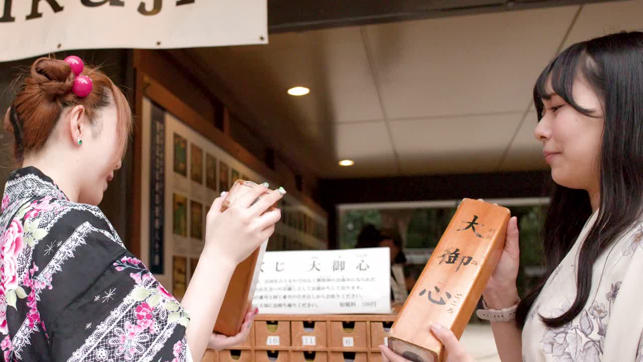 日本の神道神社で、伝統的な着物を着た2人の女性が木製のおみくじ箱を振っている。