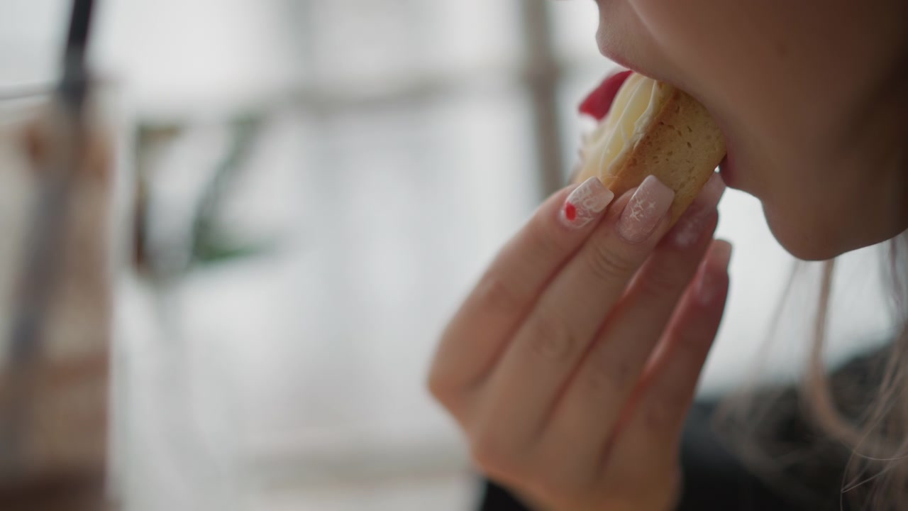 Side-view close-up of lower face and manicured hand holding small bitten pastry near mouth, showing eating gesture, visible filling, soft daylight, blurred background, warm lifestyle mood