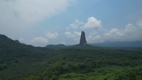 aerial view flying backwards over the forest with e pico cão grande mountain at background, in sãotome,africa
